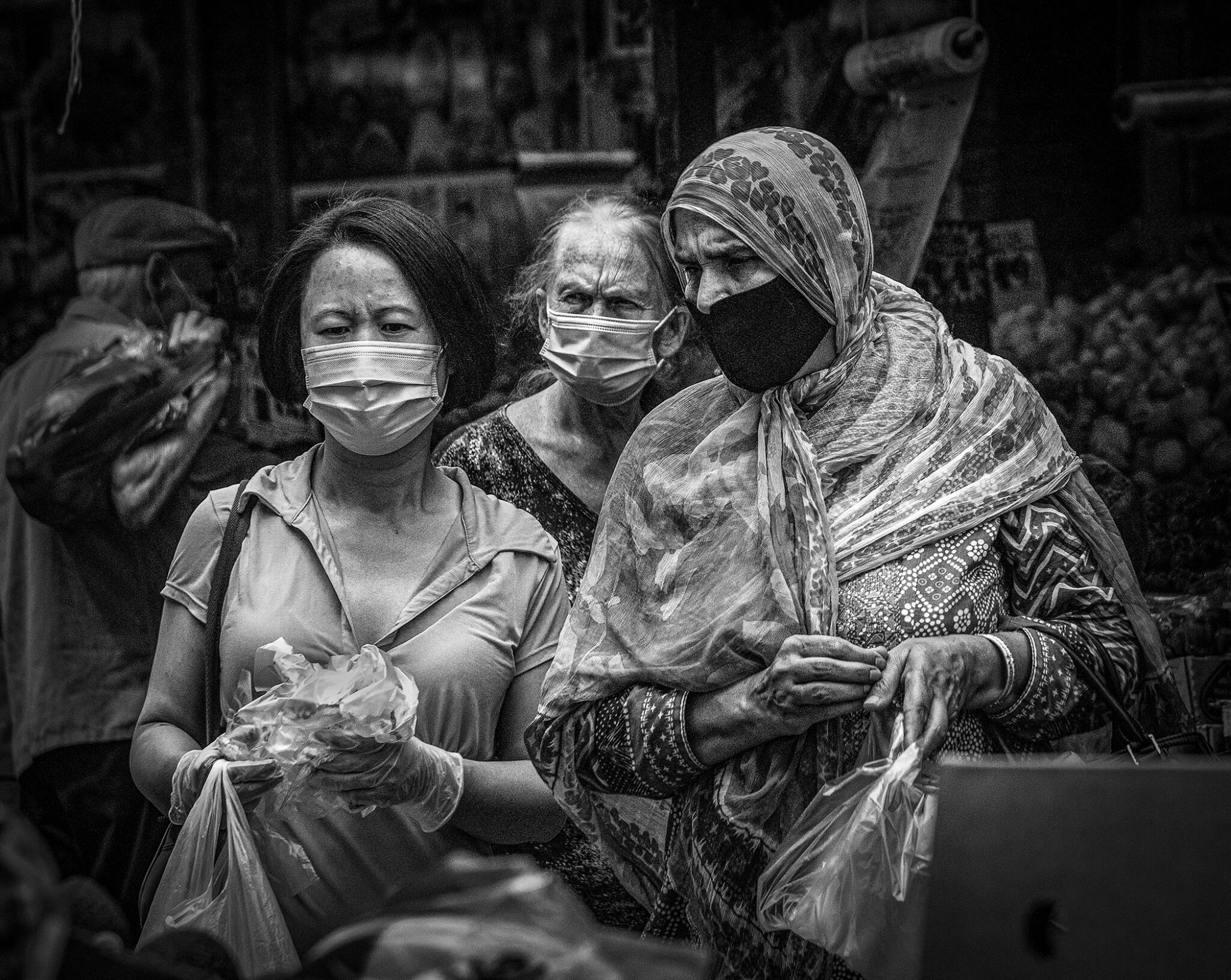 Black and white: three people with face masks squint, as one would in the bright sun, holding plastic produce bags.