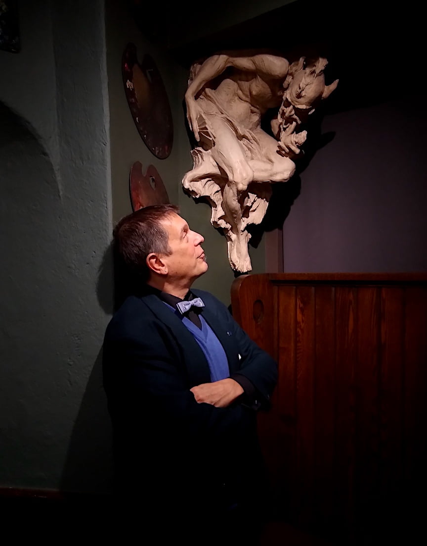 Anthony Bellov looks up at a gargoyle on the Salmagundi bar room wall close to his face. His arms are crossed, and he has a pleased expression.