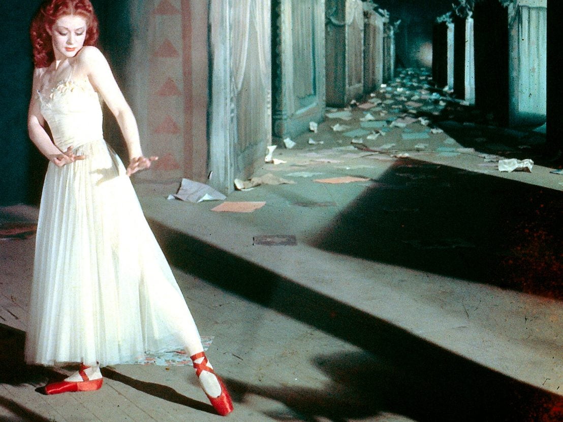 A pale, brunette woman with a white dress and red ballet slippers points her toe, her wrists bent so that her palms face the foot. She stans in front of a dirty outdoor corridor.