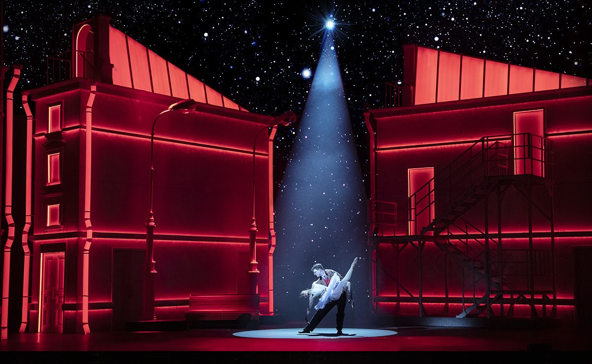 Between glowing-red set buildings on a stage are a man dipping a ballerina with her leg in the air. They are lit by a single spot light above them, the background shimmering with stars.