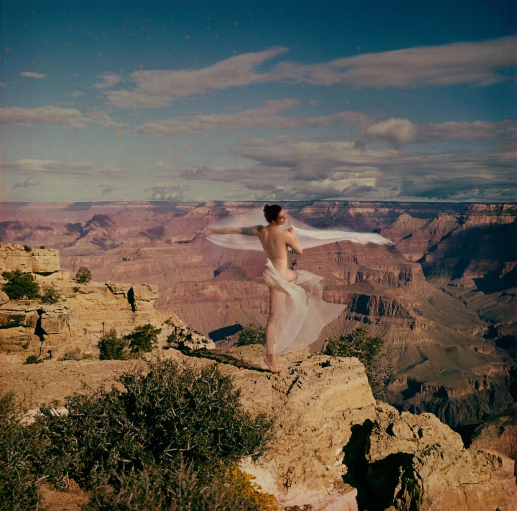 A digitally manipulated photographic piece; a nude woman stands on her tip toes, overlooking a vast landscape of canyons. Sheer white fabric is draped around her body and billows in the wind.