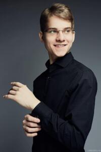 Aleksandr Bolotin is a young, sandy-haired man with glasses wearing a black button down. He adjusts his cuff and looks to the side, smiling.