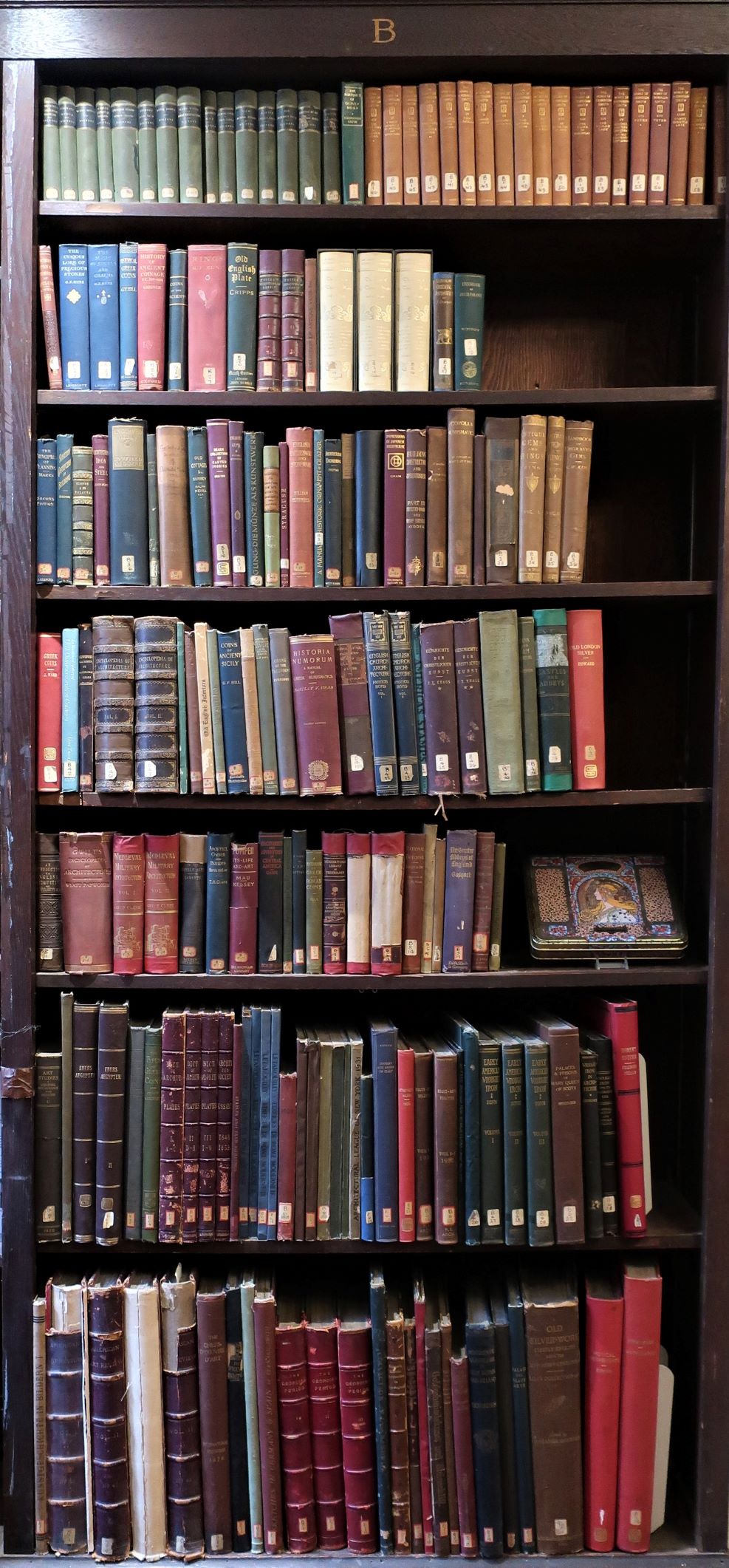 A vertical shot of a library bookshelf after some much needed dusting. Worn spines face the viewer, and a square slab featuring the profile of a woman by Alphonse Mucha is displayed on a middle shelf.