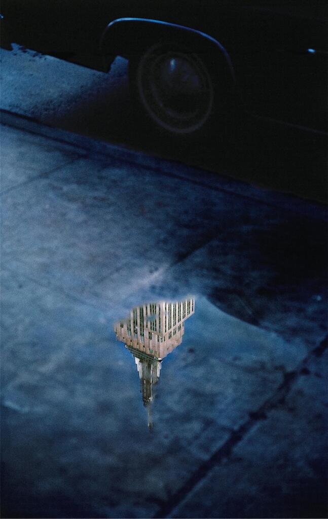 A mundane shot of a puddle by the foot of a car. Standing out from the dreary blue of the wet sidewalk, the reflection of the Empire State Building stands strong, illuminated by daylight.