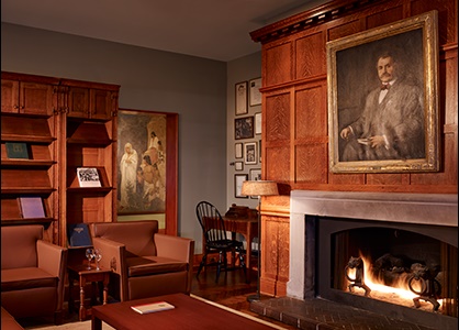 The interior of a room in the Cliff Dwellers Club: flames light up a warm fireplace. Books are displayed on shelves behind armchairs and a desk sits at the corner under an arrangement of frames.