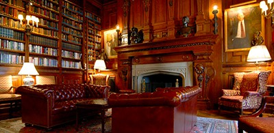 Shelves of books line the wall of the Lotus Club library. Two red chairs face each other in front of a grand mantlepiece.