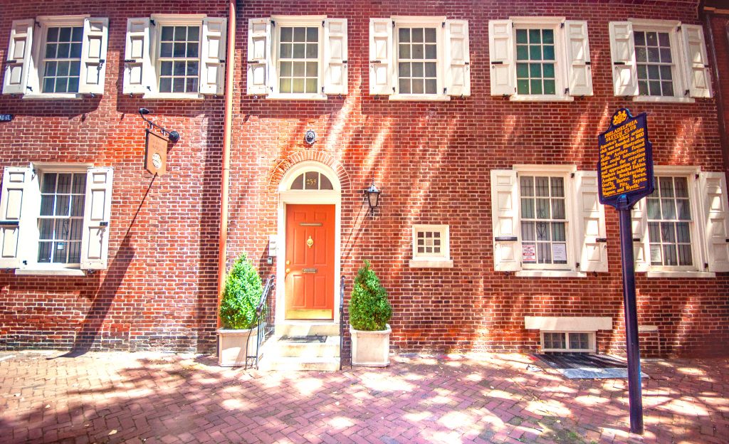 The brick building of Philadelphia Sketch Club under dappled sunlight. A row of windows line the top over a red door with potted plants on either side.