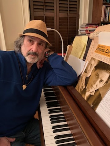 Brian Gari wears a trilby hat and a light, blue jacket. He rests his arm on the music stand of the piano he sits at so that his body opens toward the photographer.