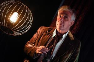 Tony Perrottet is photographed diagonally in a brown leather jacket, swirling a straw in his drink. He is lit by a spherical lamp in the foreground.