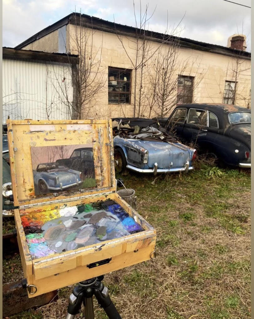 A portable easel/case hybrid is set up outside with a painting of a broken down car, juxtaposed against the actual subject.