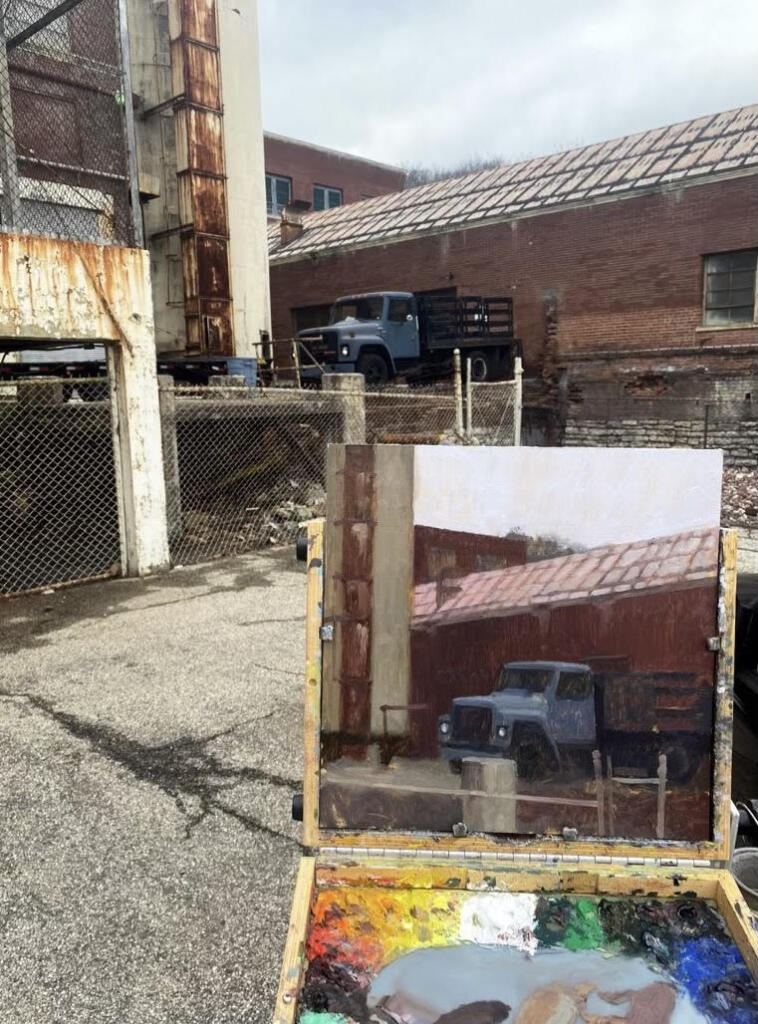 A portable easel/case hybrid is set up outside with a painting of an abandoned pickup truck, juxtaposed against the actual subject.