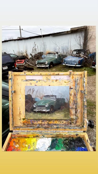 A portable easel/case hybrid is set up outside with a painting of a broken down car, juxtaposed against the actual subject among several differently colored cars.