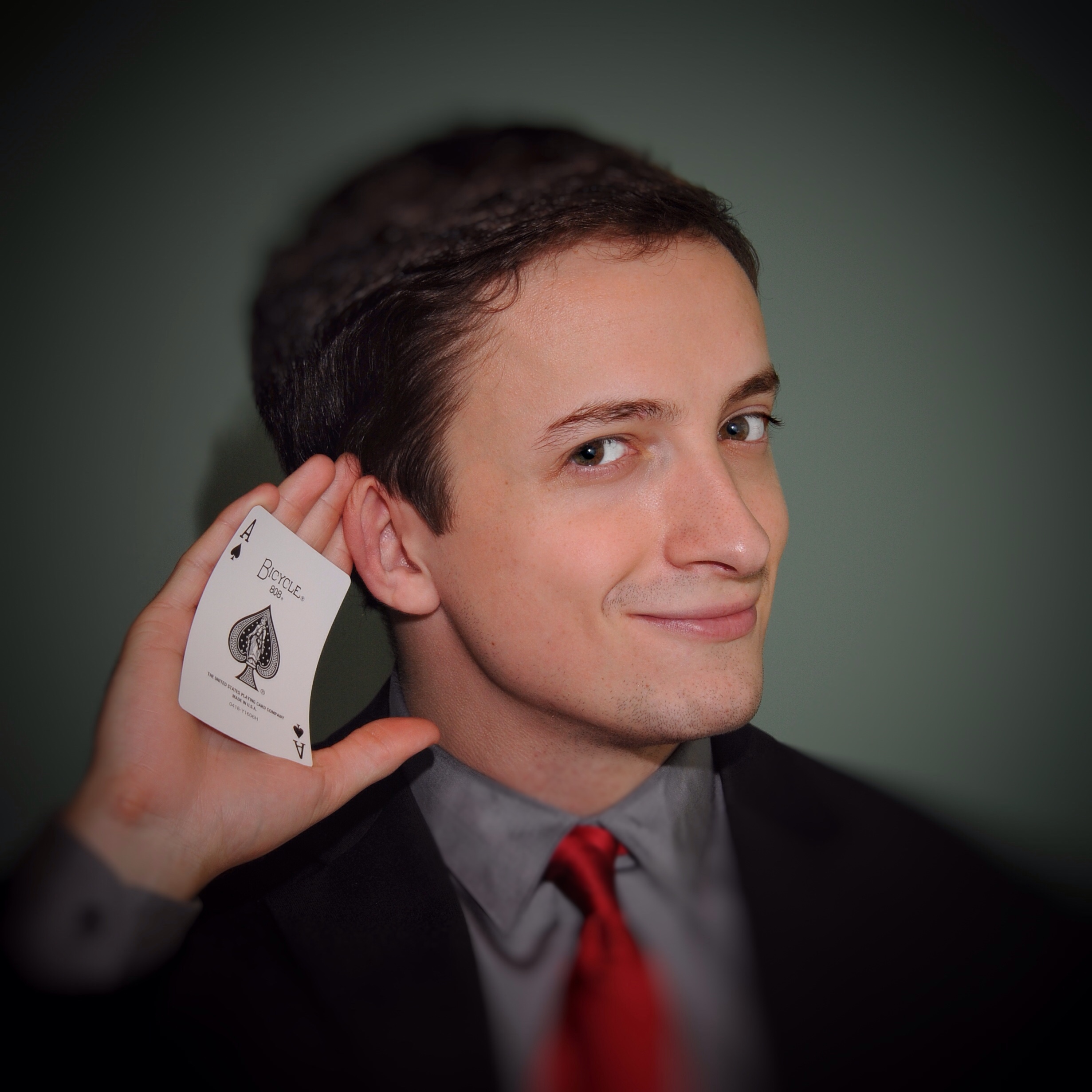 Daniel Roy is a young white man with slicked brown hair. He smirks playfully and puts his hand behind his ear with a card face up in his palm.