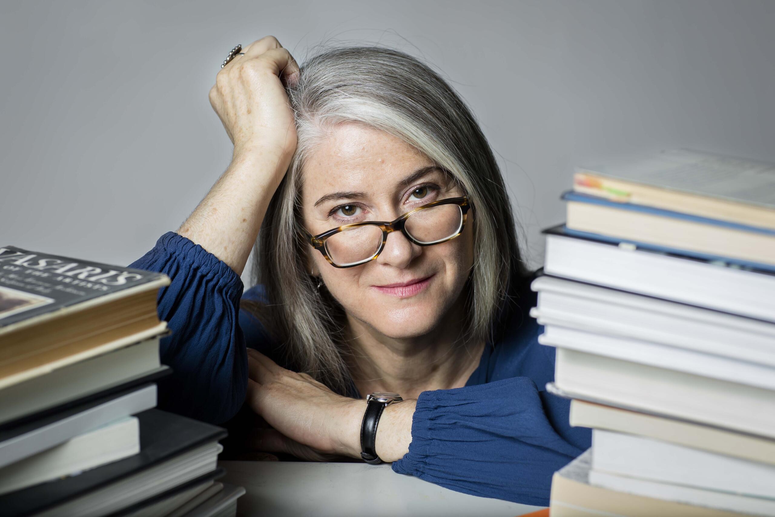 Milène J. Fernándaz is a gray-haired woman with glasses that are low on the bridge of her nose. Srests her arms on a table with stacks of books in the foreground.