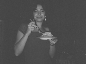 In black-and-white photo, Simona Blat smiles, putting a fork into the food on her plate that she holds with her other hand.