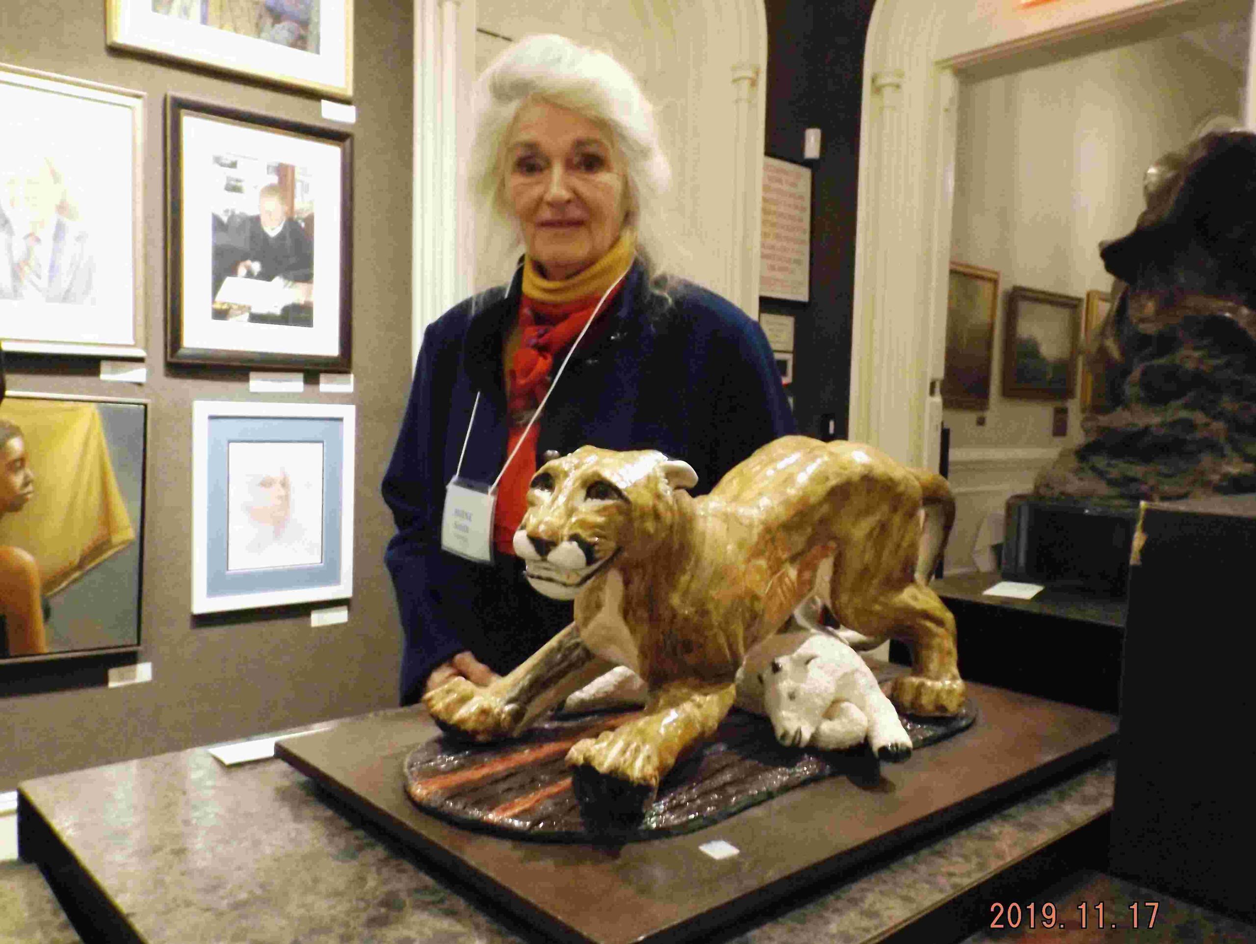A white-haired woman looks at the viewer from behind a sculpture of a wild cat, standing over a collapsed lamb.