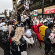 In a city, a group of people standing around a street, some in costumes, watching a woman with a rabbit mask play a guitar wrapped in tinfoil.
