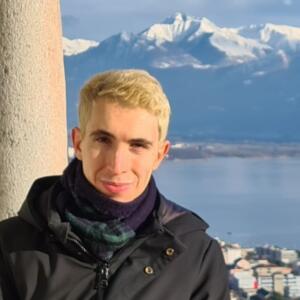 Matteo Parisi is a young man with short, bleach-blond hair. He wears a scarf and jacket and poses in front of a mountain range.