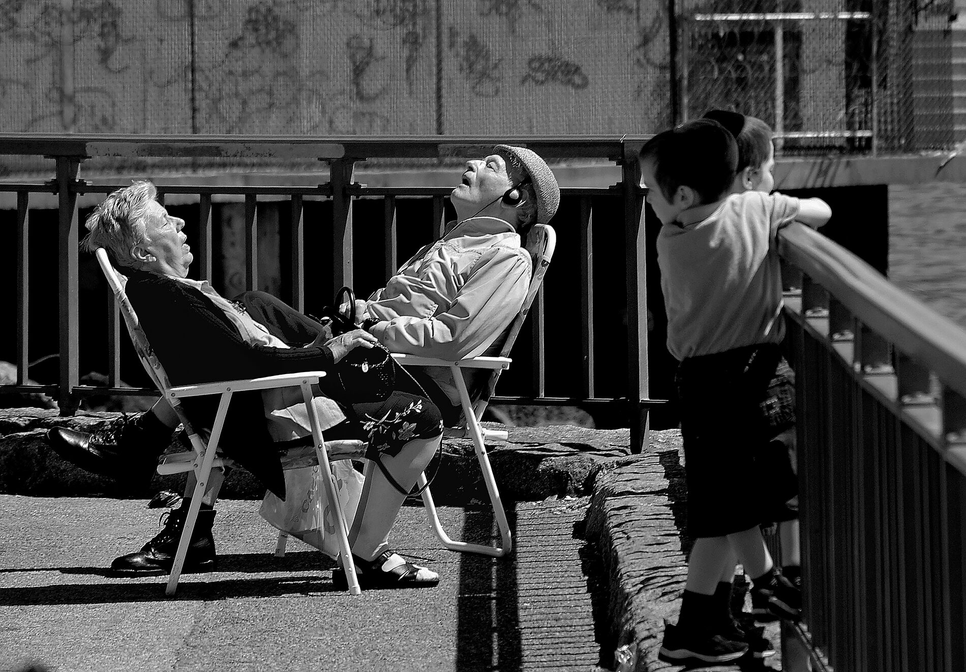 Black and white: Two little boys with yarmulkes leaning on a harbor railing look over at an old man and woman in folding chairs, one with earphones and the other clutching a purse, both conked out in a deep sleep.