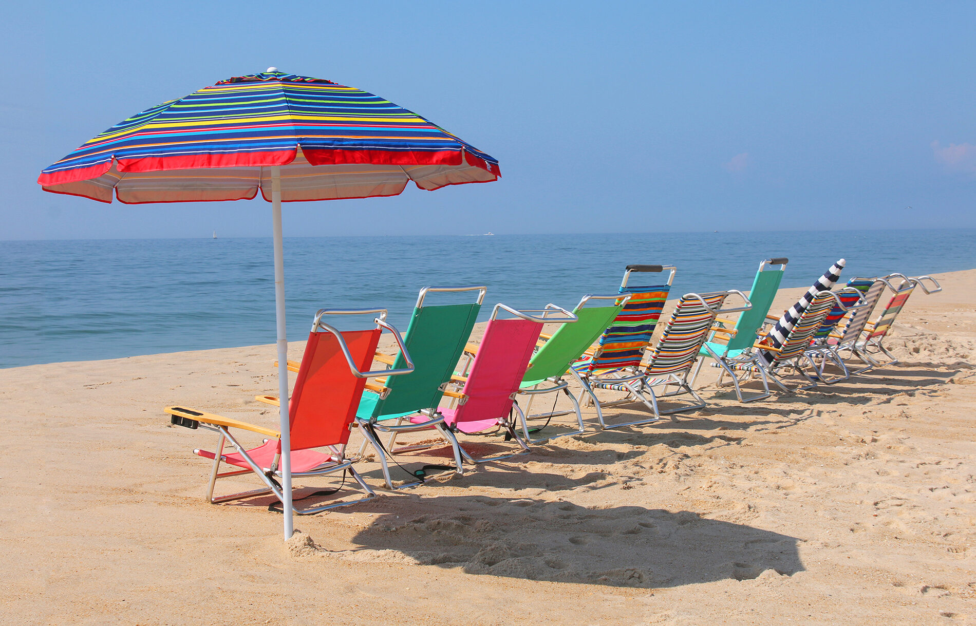 A row of beach chairs sitting on top of a sandy beach, a horizontally-striped beach umbrella standing at the end of the row.