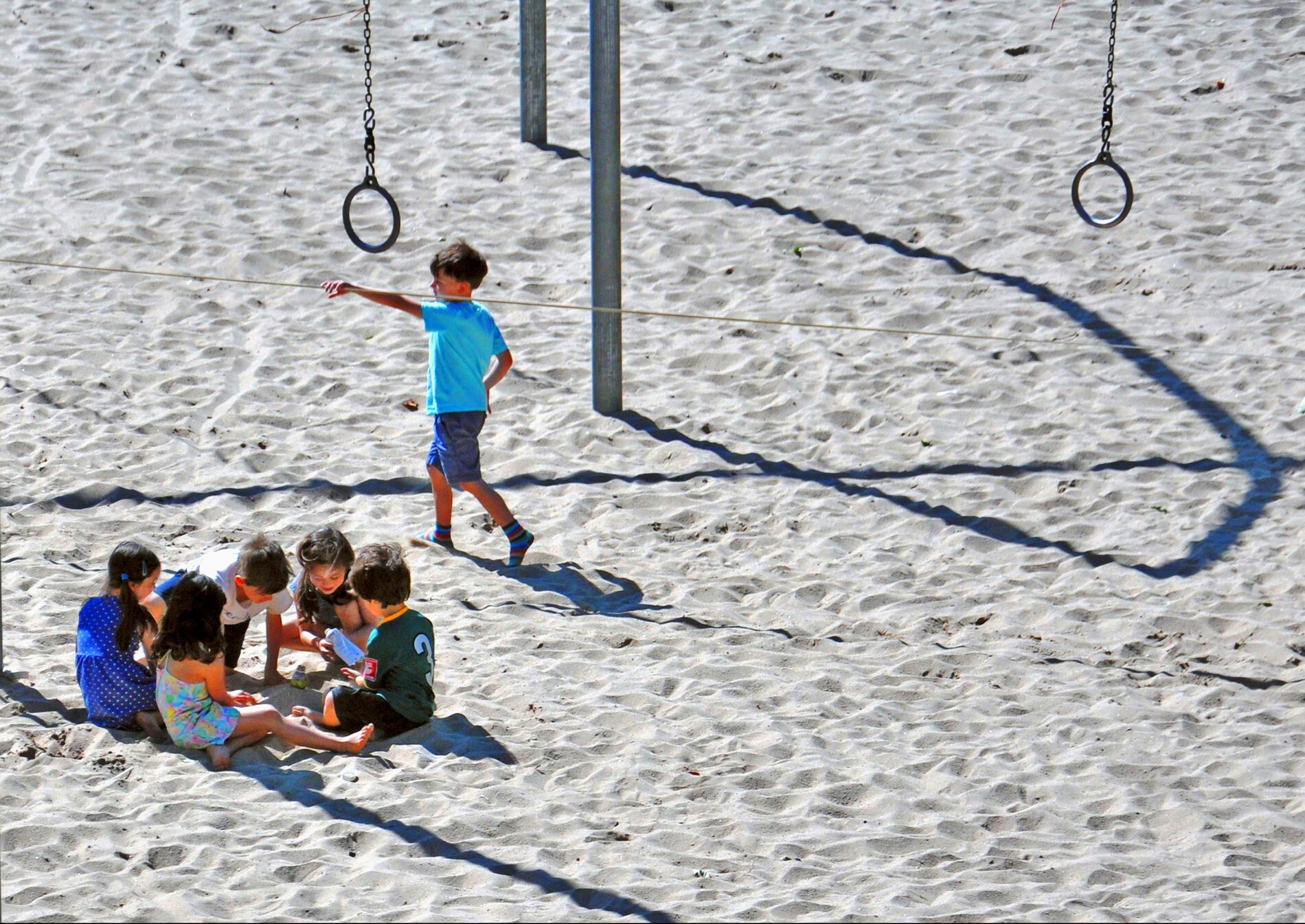 On a sandy beach, five kids sit close in a circle and another moves his hand along a long rope that is suspended four or five feet in the air. Two metal poles stick out of the sand, and two rings on chains hang down.