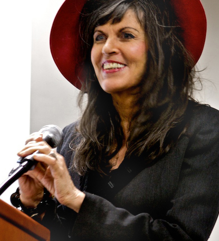 Jerelle Kraus is a middle-aged woman with dark hair and bangs. She smiles and wears a wide-brimmed had and has her and around the microphone at the lectern in front of her.