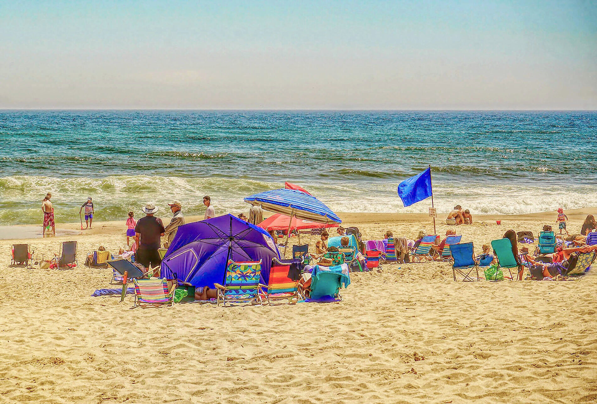 Highly saturated photo of lots people at the beach with many beach chairs and umbrellas set out.