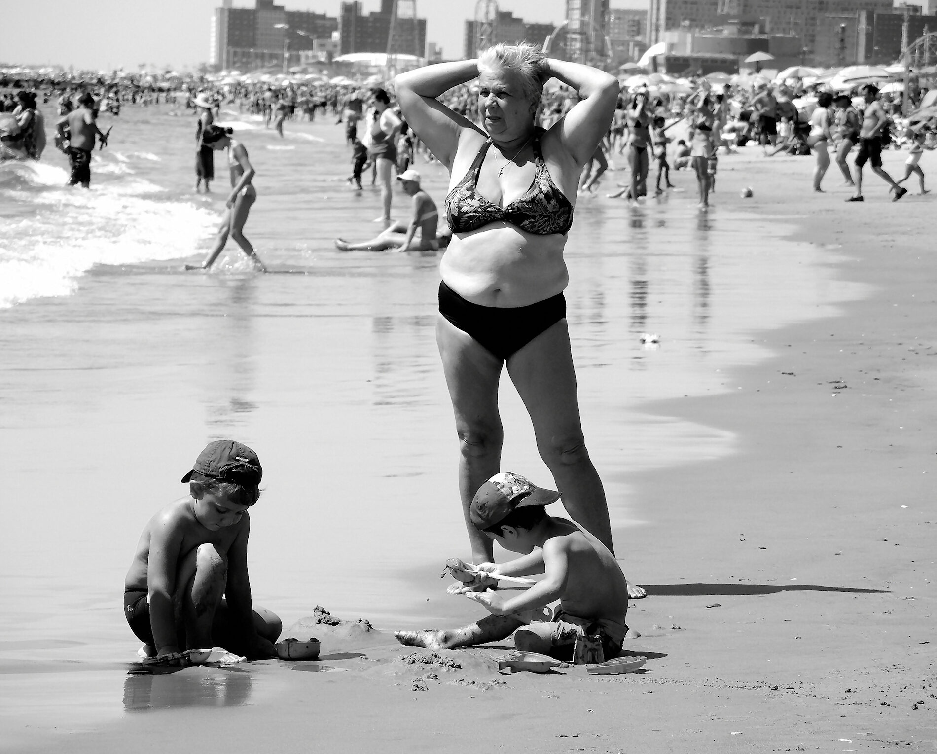 An elderly woman in a bikini standing on a crowded beach with her hands behind her head, two boys with baseball caps playing in the sand at her feet.