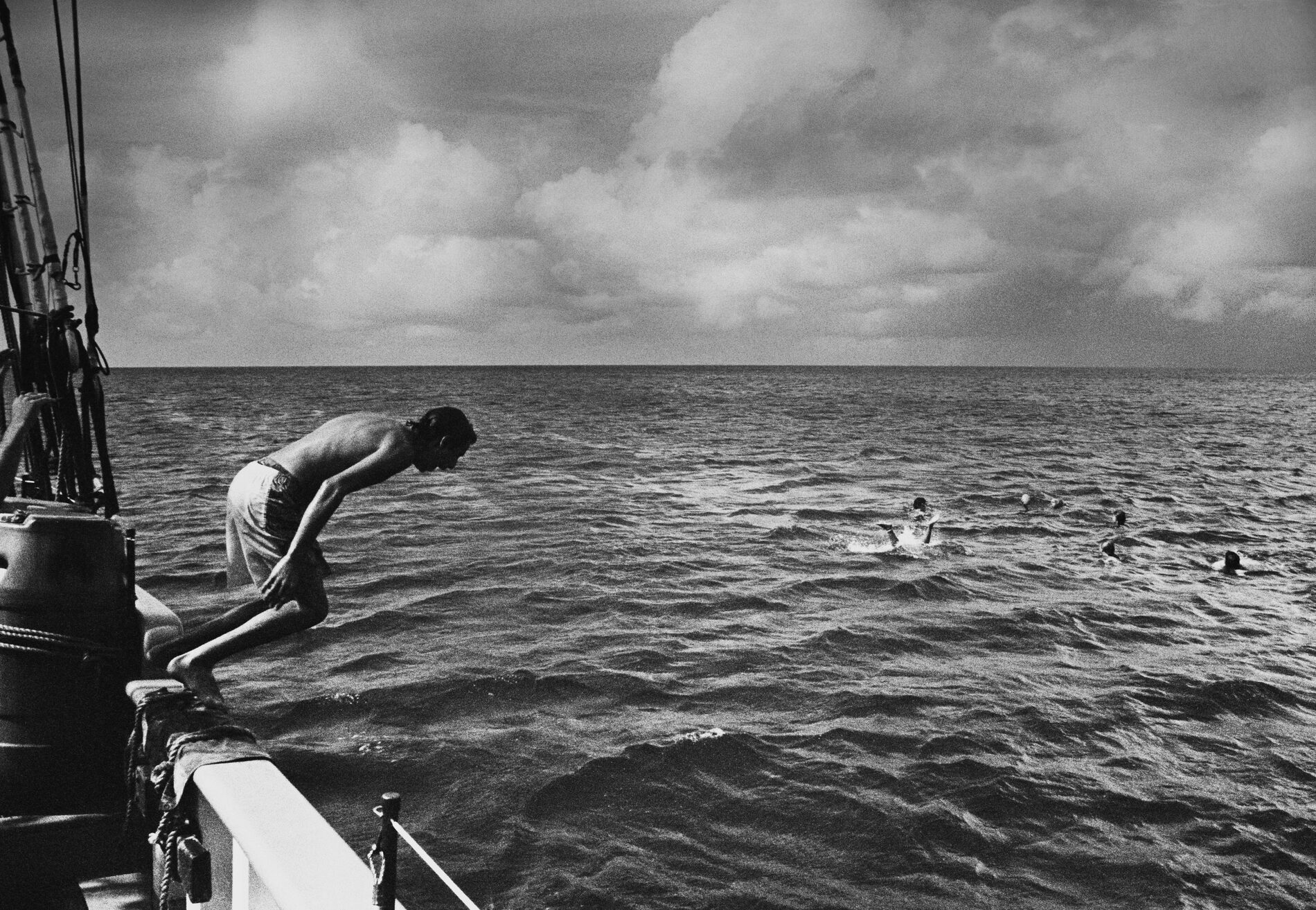 Black and white: a man in a swim suit jumps off the side of a boat. About 10 meters away, someone is in the water with their feet sticking up.
