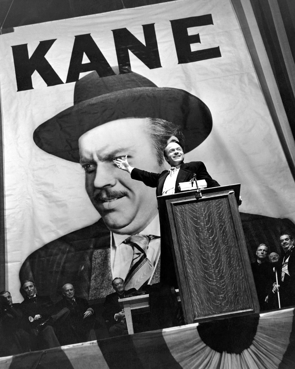 Orson Welles as Charles Kane on a stage, gestering his hand from behind a podium. Behind him are men in suits, sitting and a giant banner with his face and name.