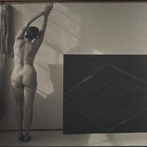 Black and white: from behind, a nude woman stretching her arms up and t the side.