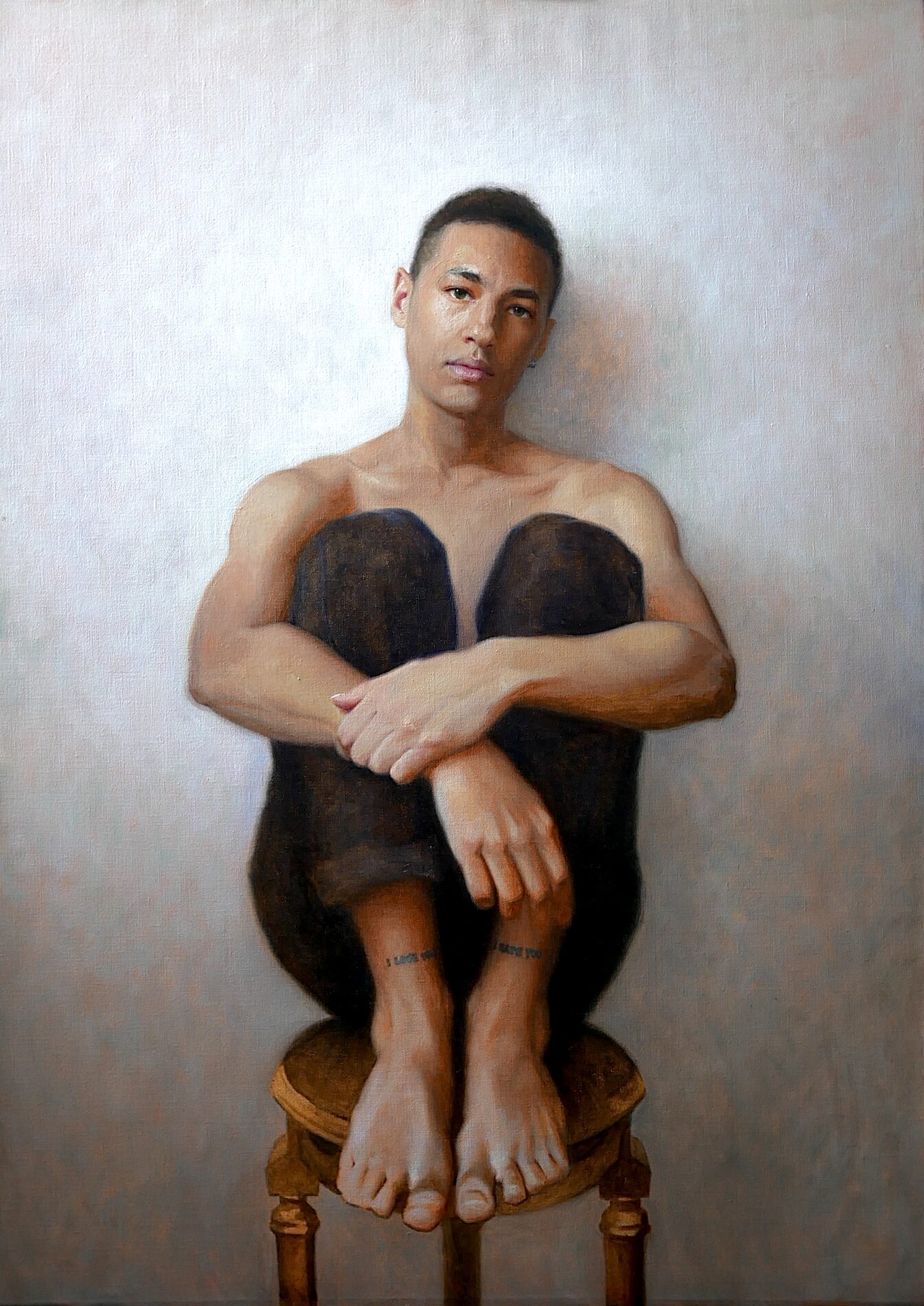Shirtless young man sitting on a stool, holding his knees to his chest.
