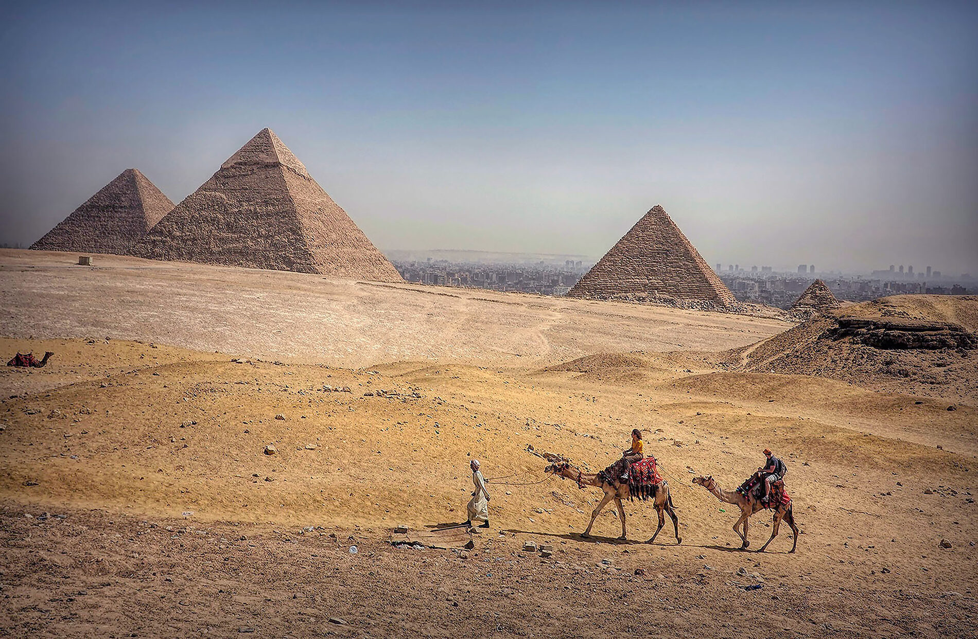 A person in a white full-body garment leading two people on camels in front of Egyptian pyramids.