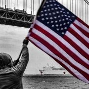 A man waving an American flag in front the New York Harbor.