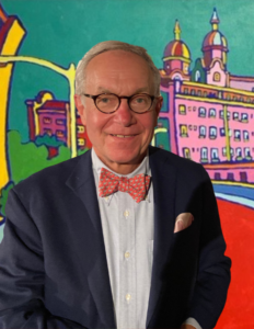 An older man in a fun red bowtie and rounded tortoiseshell glasses smiles at the viewer, in front of a colorful mural of a pink building.