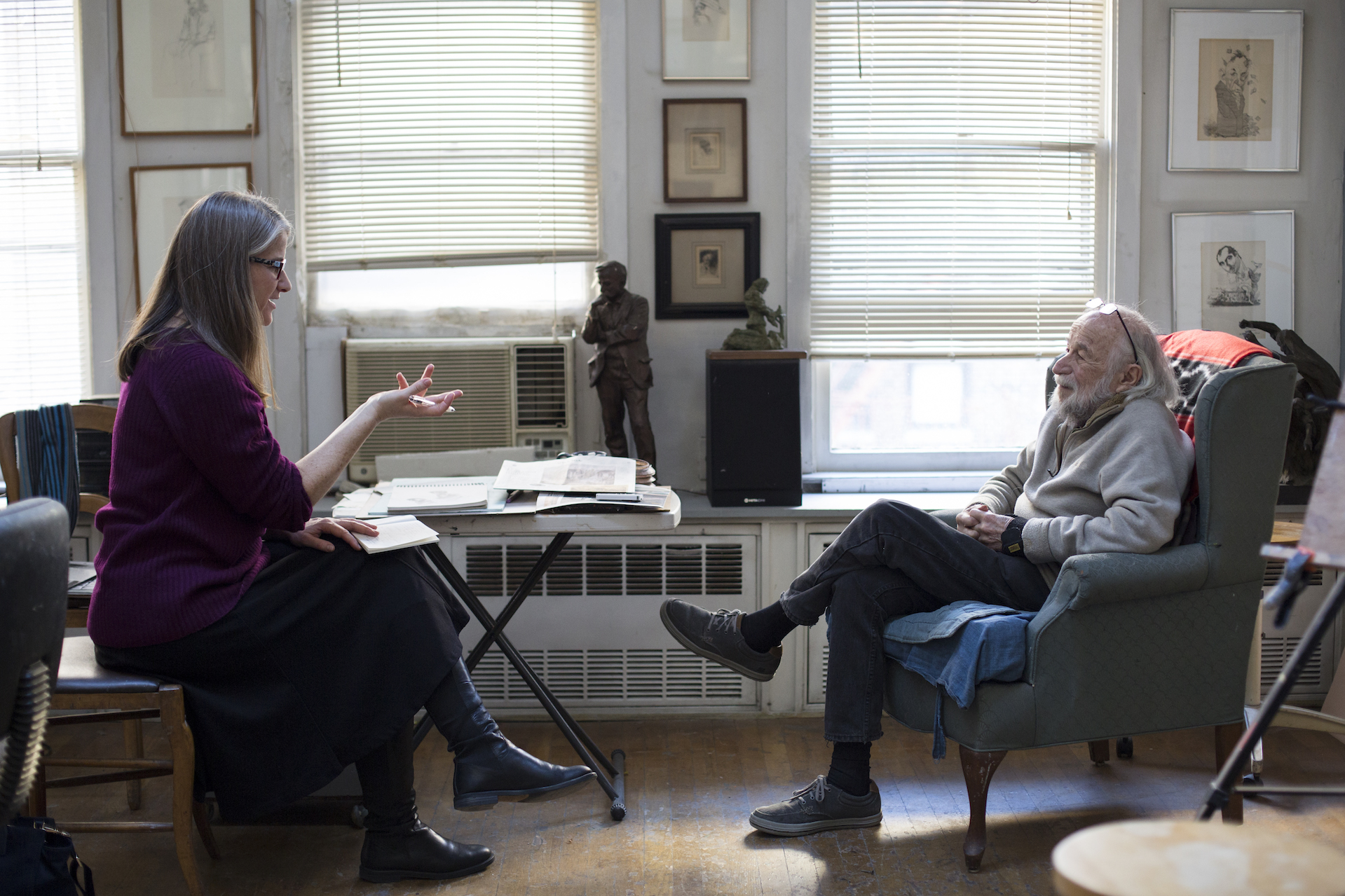 A woman engages in conversation with an older man in his New York artist studio. The walls are adorned with framed drawings.