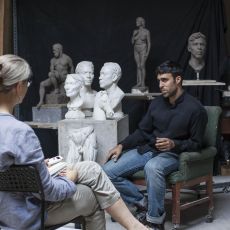 A woman is seated across from a sculptor in his studio, full of busts and full body casts.